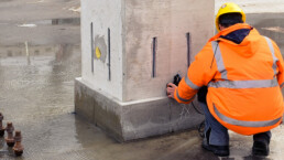 Untersuchung eines Betonpfeilers durch einen knieenden Mann mit orangener Warnjacke und gelben Bauhelm
