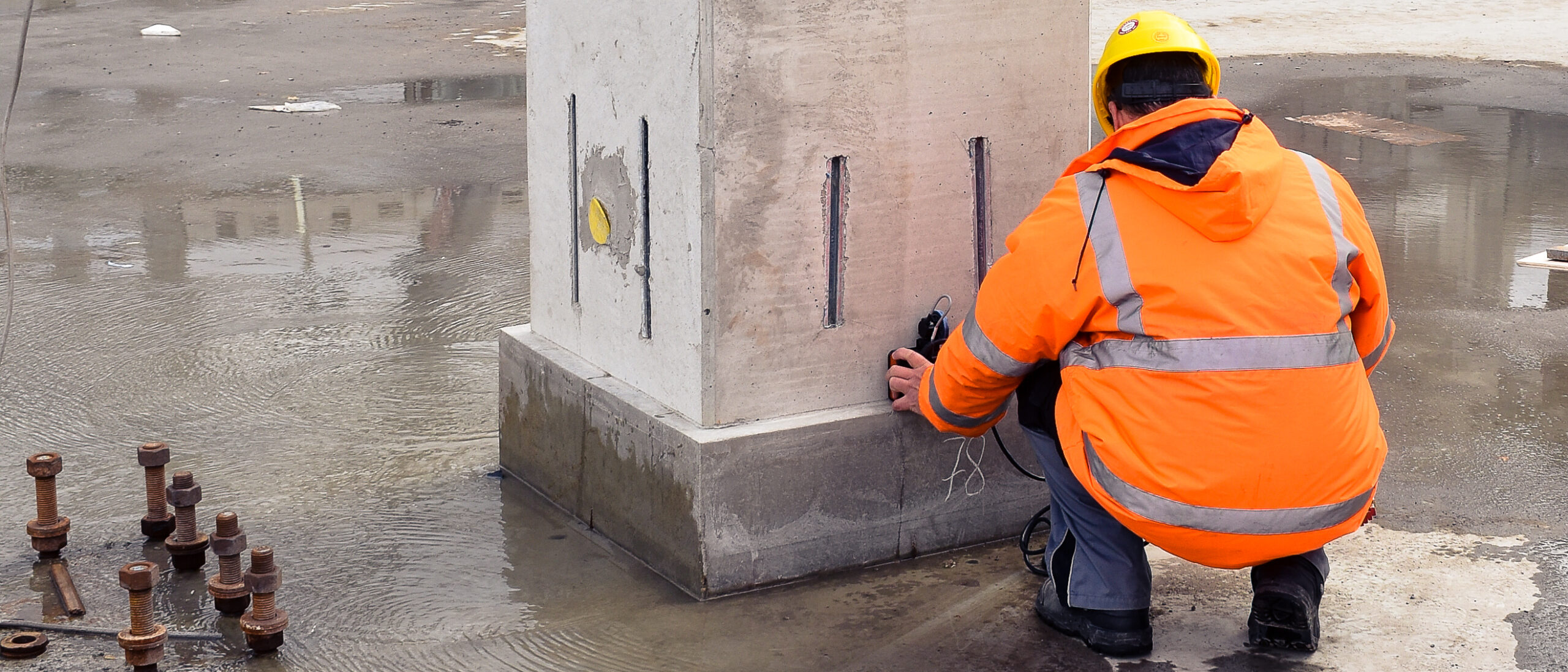 Untersuchung eines Betonpfeilers durch einen knieenden Mann mit orangener Warnjacke und gelben Bauhelm
