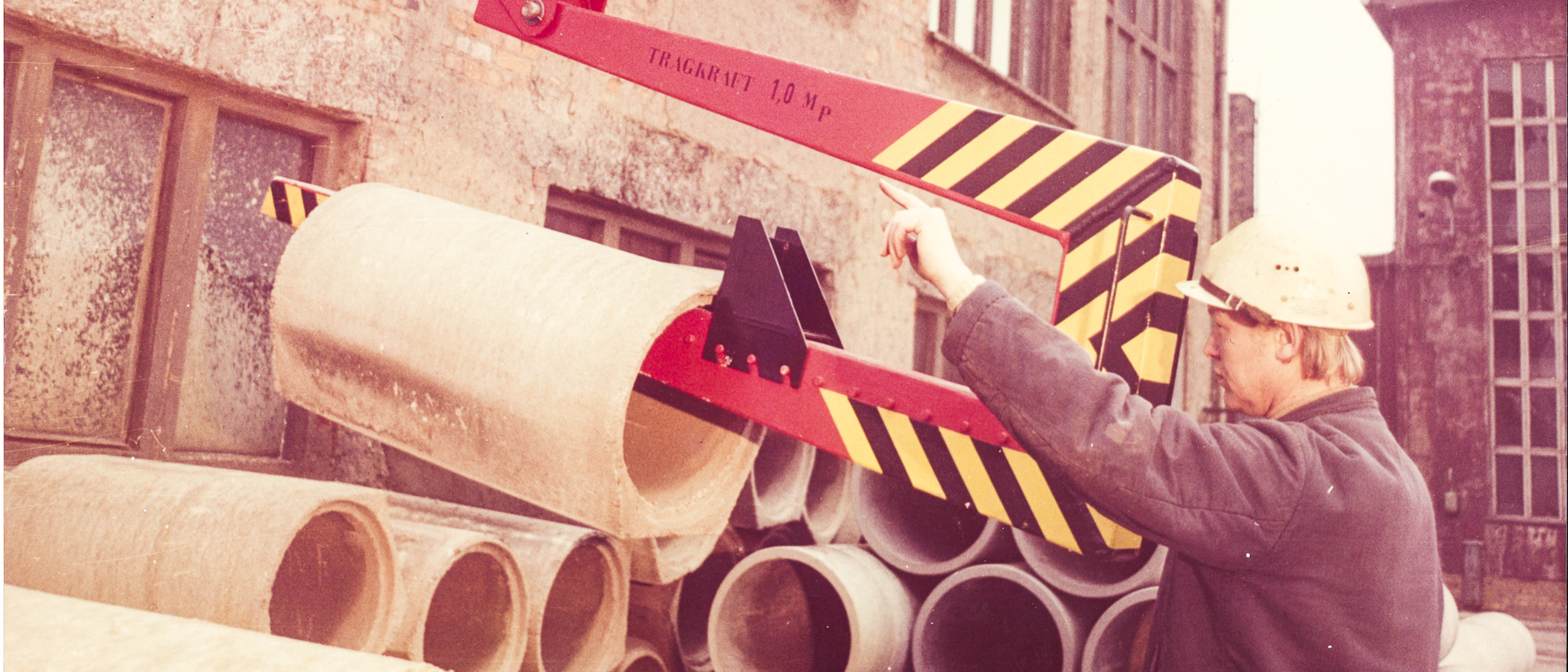 altes gelbstichiges Bild auf dem ein Mann mit Bauhelm Betonrohre mit Hilfe eines Greifarms umsetzt