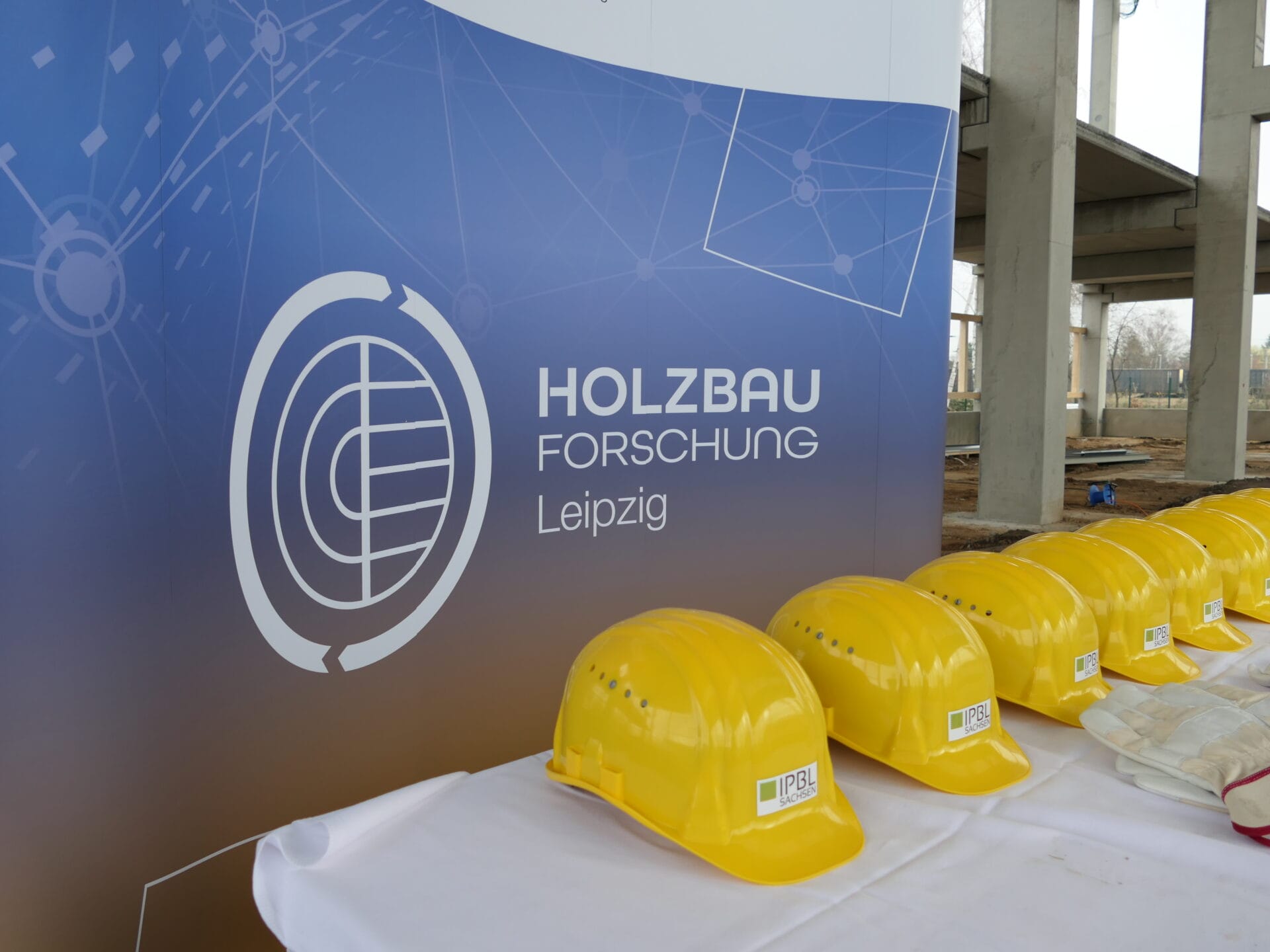 gelbe Bauhelme des InnovationsParks Bautechnik Leipzig Sachsen vor dem weißen Logo der HOLZBAU Forschung Leipzig