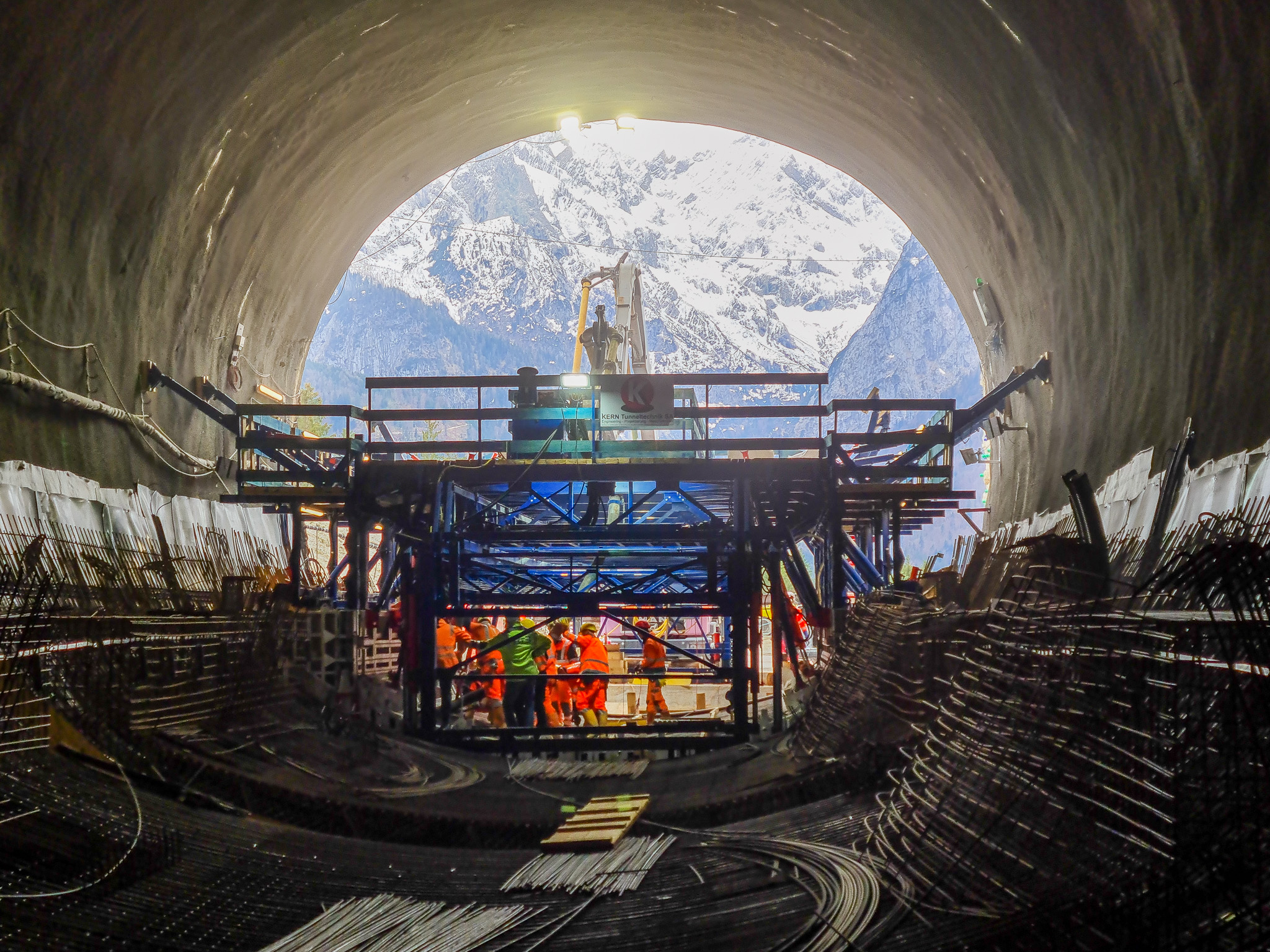 Blick aus den Ausgang einer im Bau befindlichen Tunnelröhre auf die dahinterliegenden Berge mit Bauarbeitern und Stahlbewehrung im Vordergrund